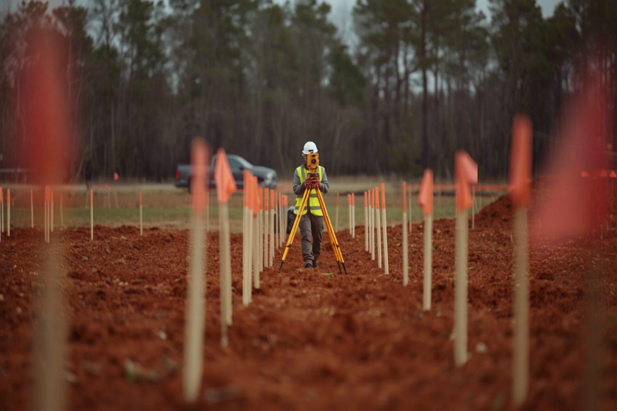 A land surveyor in a high-visibility vest and white hard hat sighting through a yellow total station on a tripod in the middle of a graded red-clay lot, framed by rows of wooden stakes with orange flagging and a pine treeline behind