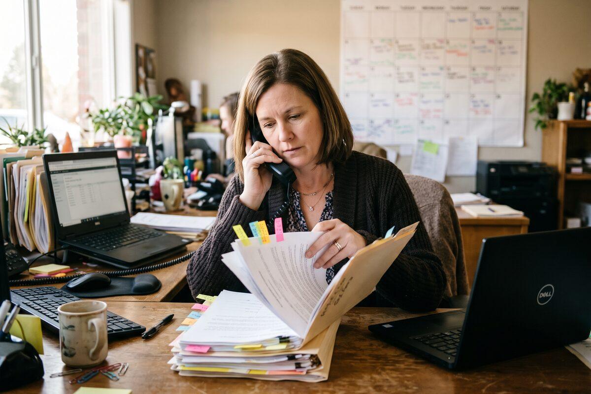 A transaction coordinator in a dark cardigan at a cluttered brokerage office desk, on the phone while flipping through a thick manila folder of contract pages marked with colored sticky flags, with two laptops, a coffee mug, and a wall calendar behind her